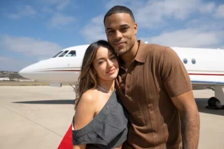 Carolina and Grant pose in front a of jet on the tarmac. Grant is wearing a brown shirt that looks a bit like a guayabera, and Carolina is wearing an off the shoulder top that looks like the waistband of some trousers.