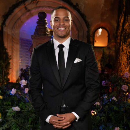 Grant poses in front of the McMansion. He has medium brown skin and is wearing a black shit and tie with a white pocket square. His hands are folded in front of him and he's smiling at the camera. He has a very nice smile.