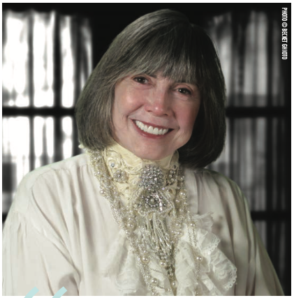 Anne Rice, a white lady with a grey and brown shoulder length bob with bangs, is wearing a VERY PUFFY SHIRT with some kind of neck piece that's silver and iridescent beads looped into a sort of bead cravat 