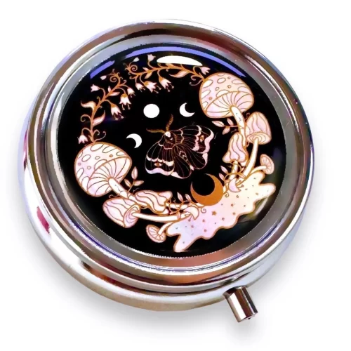 A silver metal pillcase that is round with an image of mushrooms and a moth against a black background