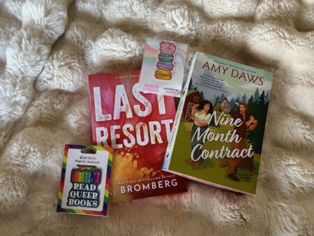 Two books and two bookmarks on a white blanket. The bookmarks are a stack of macaron, and a tile that reads READ QUEER BOOKS. 