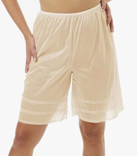 A pair of beige loose diaphanous shorts worn by a white person with one hand on their thigh and the other on their hip. The shorts are nearly knee length but have three stripes of lace trim which are the points to which you can trim them to be shorter
