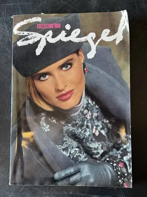 the Spiegel 1989 catalog featuring a white woman with a blonde bob in all grey - jacket, hat, printed blouse, and leather gloves. she's leaning into the camera looking very serious