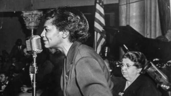 Jones looks intense as she leans forward to speak into a standing microphone, plainly dressed with her hair in a tight bun. Elizabeth Gurley Flyn, an older white woman in glasses, sits behind her.