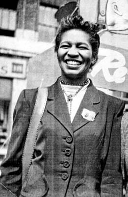 A balck and white photo of Claudia Jones, probably in her twenties, facing the camera with a huge smile, a bag over her shoulder and some kind of badge pinned to her lapel