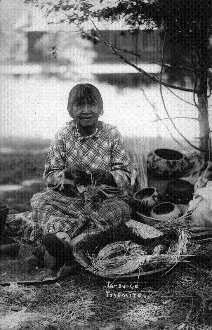 Ta-Bu-ce in black and white photo. She wears a checkered dress. She is smiling at the camera and demonstrating basket weaving.