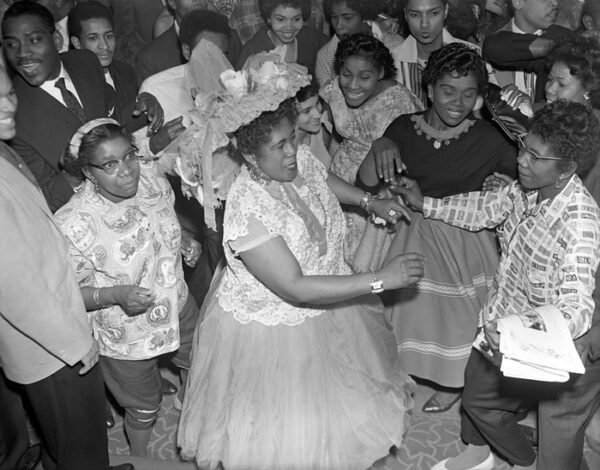 A black and white photo from the first carnival shows a crowd dressed up with, at center, a middle-aged Black woman in a white lace blouse, long gauzy skirt, and hat adorned with flowers and ribbons. Everyone is dancing and smiling.