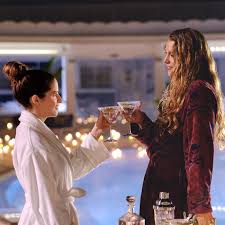 Stephanie and Emily in bathrobes toast each other with martinis poolside