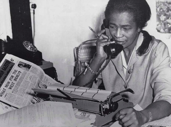 Jones in her London office at a desk that is covered with papers including an open newpaper. She is on the phone, pen in her fingers, in front of a typwriter.