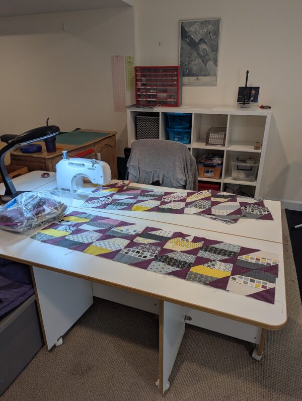 my sewing area: a white sewing table with a fold-down leaf, a chair, and some ikea kallax shelves with bins, drawers, and storage containers on shelves containing diferent sized fabric. There's a red hardware organizer on top, which is also filled with scrap squares and my sewing tools There's also a dresser to the left with a cutting mat on top.