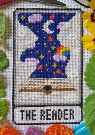 A cross stitch of a tarot card called The Reader, with a book open and a deep blue sky with rainbows, stars hearts and moons rising from the pages 