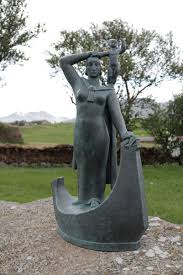 color photo of a grey statue that depict Gudrid standing in a viking boat and holding up her infant son.