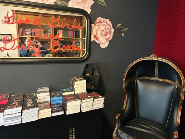 Alt: A book display against black and merlot-colored walls with a black and gold gilt chair. A mirror reads in lipstick-looking paint Good Girls Read Dirty Books.