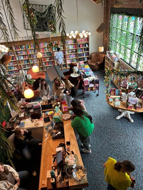 Alt: A cozy bookshop with half a dozen people, a big sunny window, and fake greenery draping from the ceiling