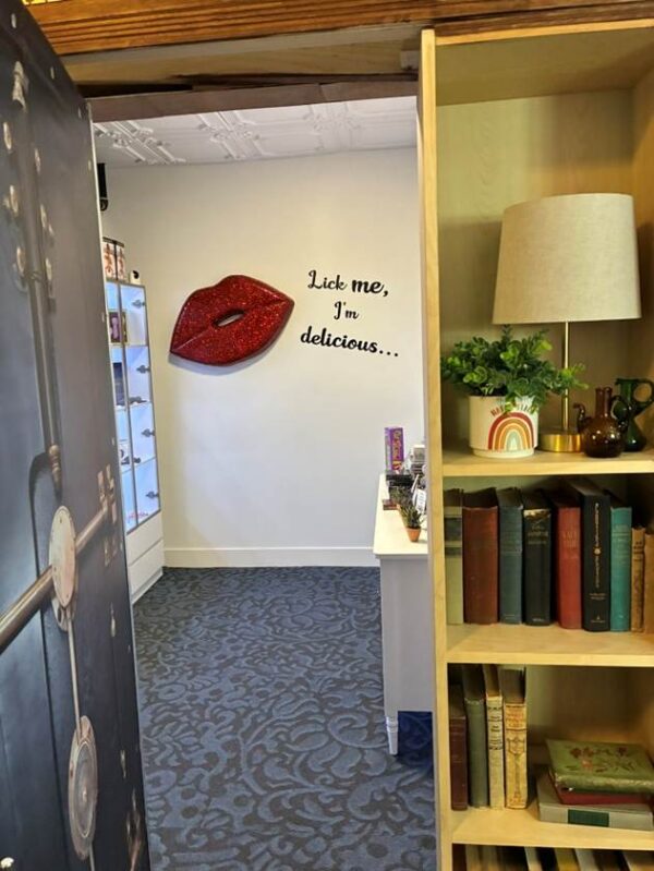 The bookshelf has swung open to reveal a white room with giant red glittery lips on the wall and the words, Lick me, I’m delicious.