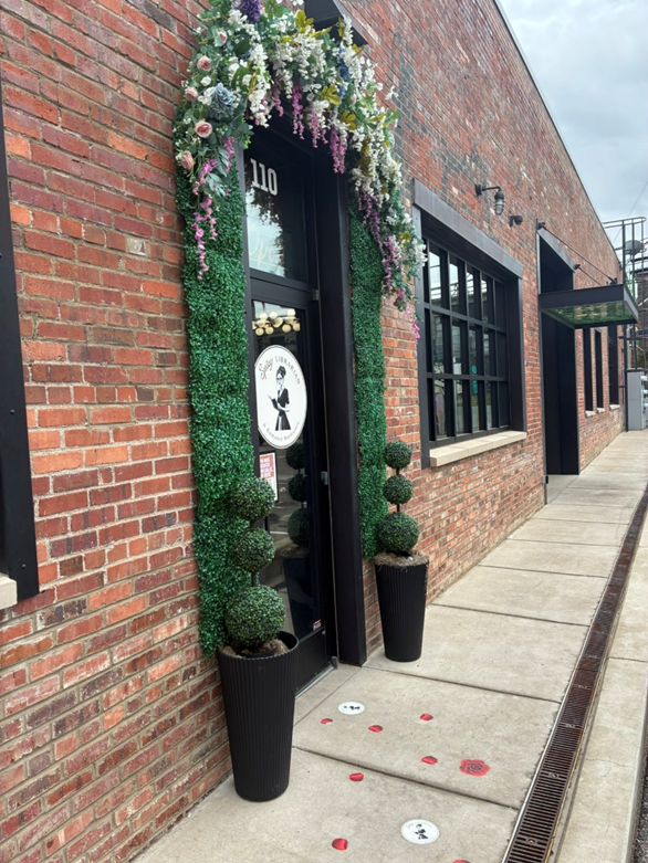 Alt text: A brick building with a door that says The Spicy Librarian. Around the door is an archway of fake flowers and greenery.