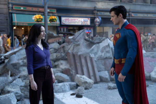 Rachel Brosnahan as Lois Lane in a purple vneck blouse and black pants standing in front of some massive concrete rubble with Superman, in costume, smiling at her 