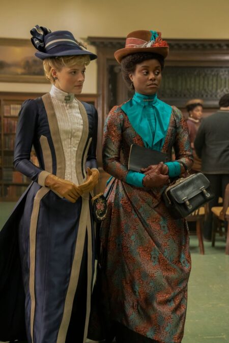 Marian and Peggy stand next to each other. Marian is in a blue gown and hat trimmed in gold while Peggy is wearing a blue and orange patterned gown with a blue blouse and hat