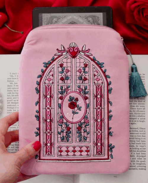A pink zipper bag with a tassel on the zipper with embroidery on the front that shows roses stained glass and an arch of leaves 