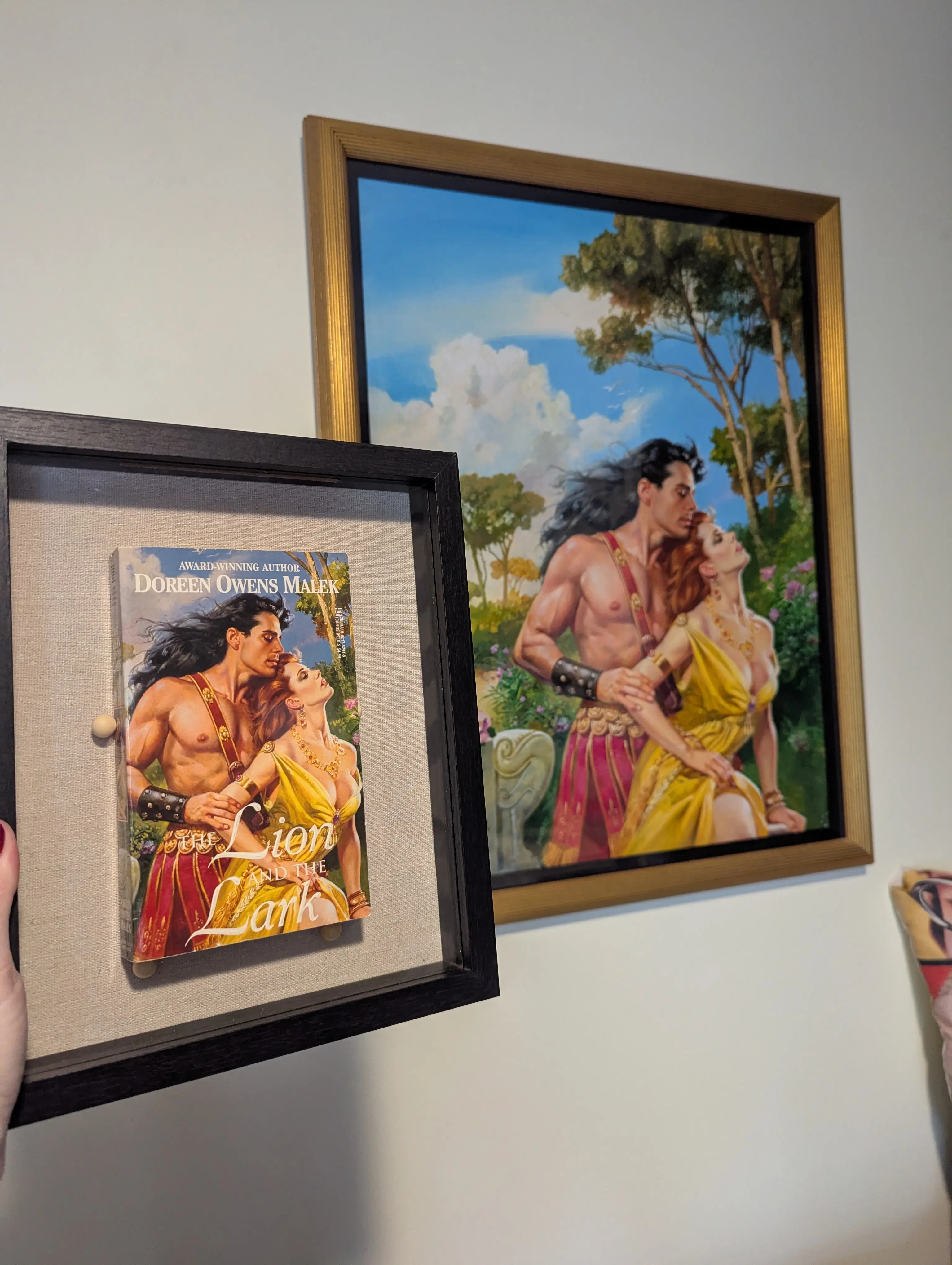 A picture of my hand holding a shadowbox with a copy of Lion and the Lark, with the oil painting of the cover on my office wall. John DeSalvo is in a hot pink gladiator toga and a woman with impossibly uplifted breasts is arching her back against him
