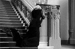 In a black and white scene, a woman dressed in a black gown faints very gracefully.