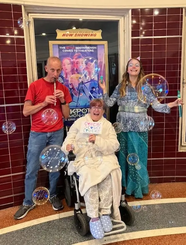 Carrie, Glen and their daughter at K Pop Sing along. Glen is wearing a red tshirt and jeans and is holding a recorder, Linden is wearing a teal costume, and Carrie is in a power wheelchair in a bathrobe, slippers and is holding a sign that says COUCH COUCH COUCH