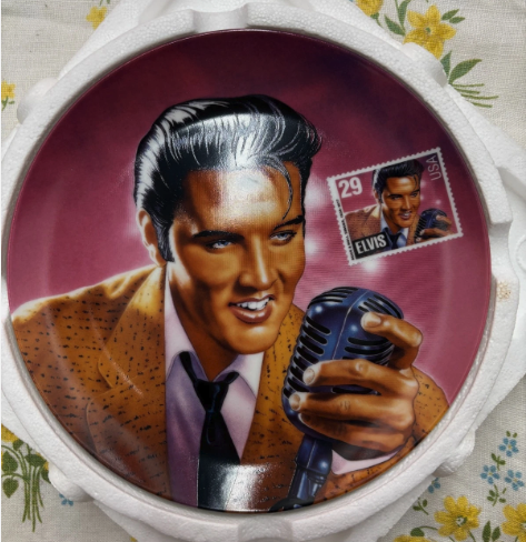 A commemorative plate with Elvis Presley and an image of his tribute stamp from the USPS the background is pink and the illustration is 50s pompadour elvis singing into a mic