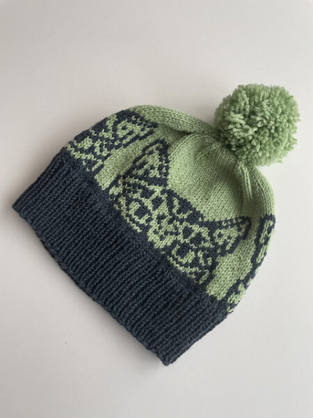 A green hat with a black brim. Black colorwork makes a cat face appear peeking over the brim.
