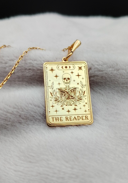 A gold tarot card miniature in gold with a picture of a skeleton reading a book with the caption THE READER. it's on a fuzzy background with a thin chain