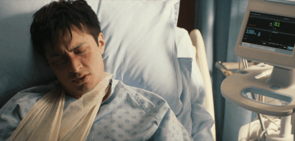 Shane looking uncomfortable in a hospital bed with a sling and gown on, with a monitor next to him reading 81 bpm