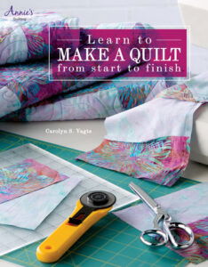 Learn how to make a quilt from start to finish by Carolyn Vagts, with a pasel batik fabric and pale white blue fabric pieces inthe front with quilting tools and a folded quilt in the back using the same fabrics