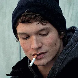 Ilya, a cigarette dangling out of his mouth, in a beanie and a coat, looks up at Shane from the legs slowly up to his face