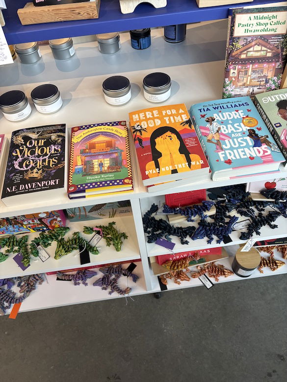 A close up of one of the tables showing Our Vicious Oaths by NE Davenport, Here for a Good Time by Pyae Thet War, and Tia Williams' Audre and Bash are just friends. Below are shelves with 3d-printed articulated baby dragons in green, purple, orange, and lavender