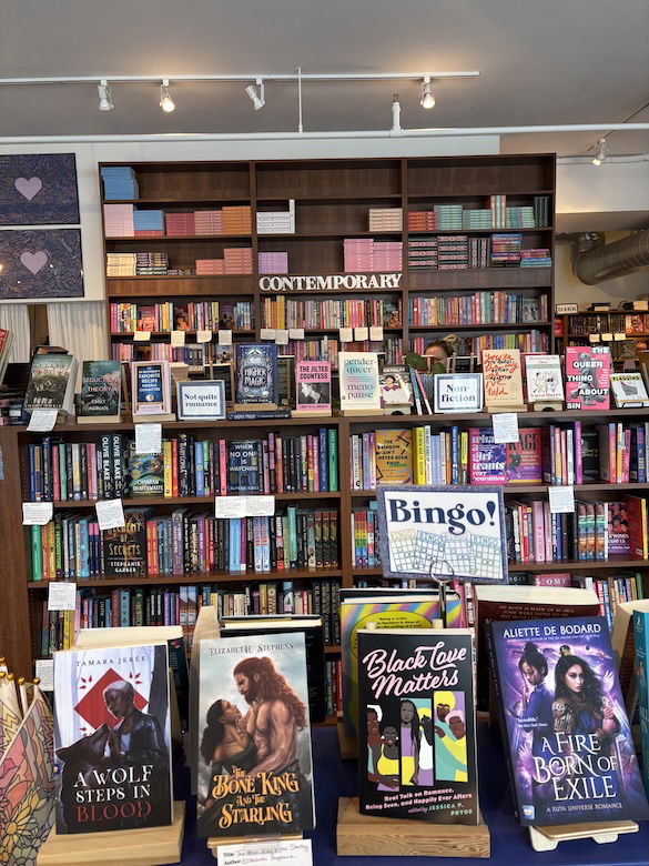A view of the store from one wall: in front are books on a table with a sign that says BINGO! and books that include Black Love Matters, The Bone King and the Starling, and a Fire Born of Exile. Behind the table are bookshelves for Not Quite Romance and Non Fiction, and against the far wall is Contemporary romance