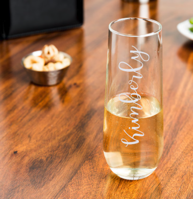 A stemless champagne glass with a name engraved on the side, sitting on a rich wood surface with a tiny and blurry bowl of nuts in the background