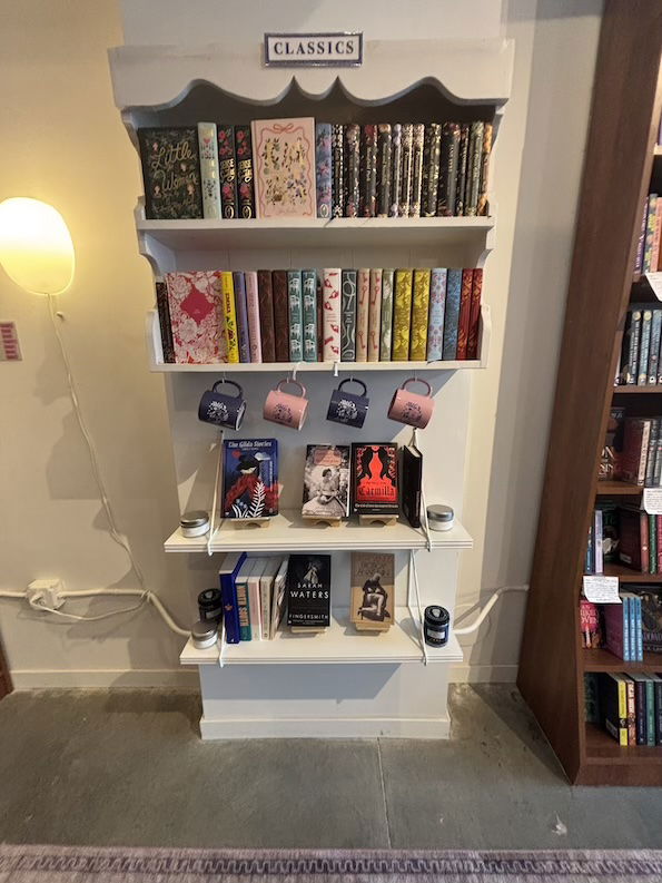 The Classics display: ornate bound editions of classic books on a narrow shelf with mugs for sale hanging below the middle shelf. Titles visible include Fingersmith by Sarah Waters, Little Women and Carmilla