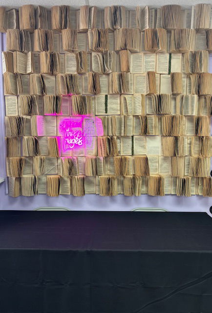A wall of open books stapled to the wall with a neon sign tucked into the pages. One of the neon words is pages but I can't make out the rest