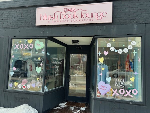 The storefront of the Blush Book Lounge, with a pink sign on a dark grey building with lit windows showing books and painted deocrations