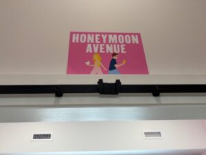 A pink sign with figures from an Emily Henry book walking away from each other reads HONEYMOON AVENUE