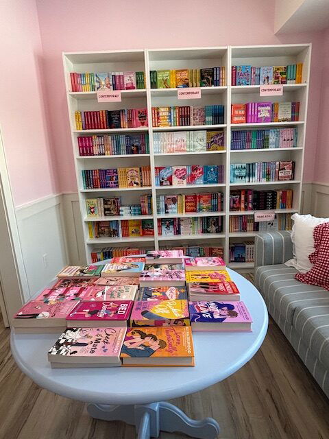A pink wall with a stuffed bookshelf in front of it. On a round table are many books waiting to be browsed, and there's a sofa to the side
