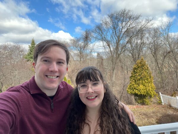 Brian and Amanda outside overlooking some woods and a small evergreen. They're smiling at the camera 