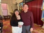 Amanda and Brian standing in a civic office. Amanda is a petite white woman with long brown wavy hair, and is holding a white bouquet. She's wearing a black top and white trousers. Brian is a white enby wearing a burgundy shirt and grey trousers