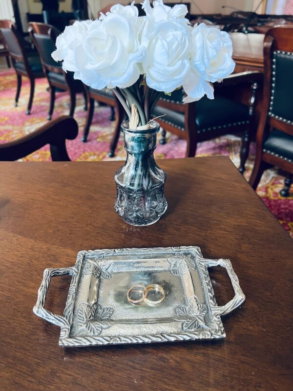 A photo of their wedding rings on a rectangular silver tray with a bouquet of white flowers in a vase behind it on a wood surface 