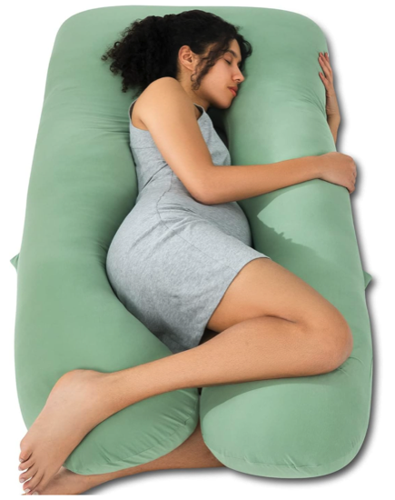 A green body pillow that is u shaped with two long arms. a woman who is pregnant is sleeping on her side with one leg and her arms around one of the arms of the pillow