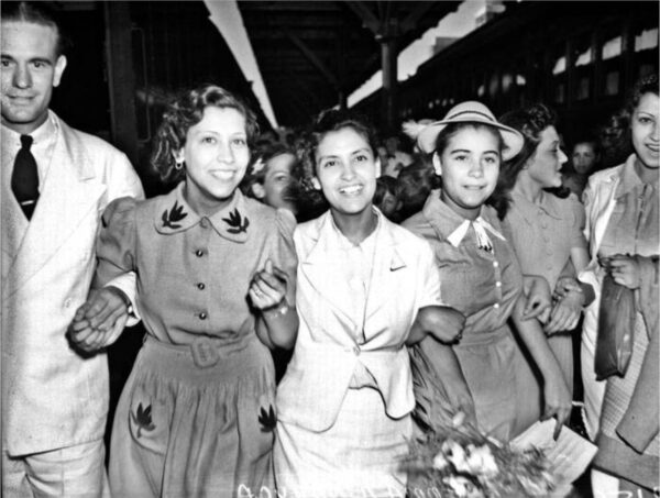 black and white photo of people standing arm in arm, Emma Tenayuca is center wearing a white suit. The two women on either side of her are wearing dresses that render grey in the photo, so Emma stands out. They are all smiling broadly 