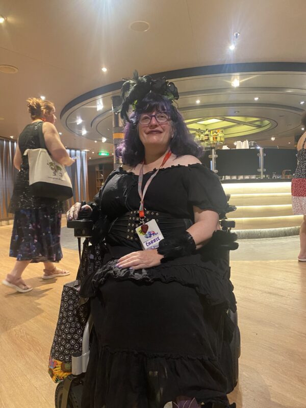 CarrieS (short white woman) with blue and purple hair, wearing a headress with a fake crow on it and a long black dress