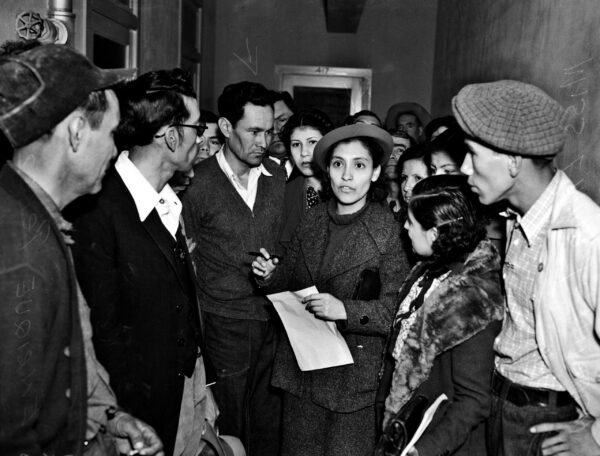 black and white photo shows Emma in the center of a crowd of men. She is wearing a hat and a coat and is holding a white paper and pen in her hand. It appears she is telling them something as they are all looking to her, and she is the center of their attention and the photograph