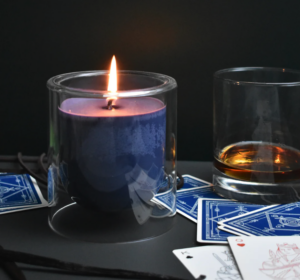A picture of the Scoundrelwood candle, a deep blue violet candle in a glass container, with playing cards and a tumbler of brandy alongside