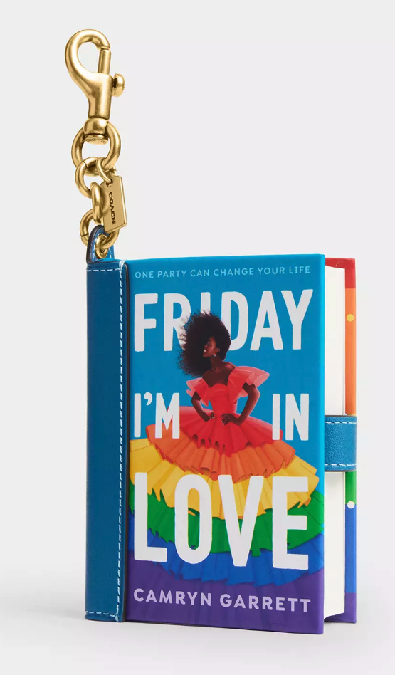 A book charm of Friday I'm in Love by Camryn Garrett, featuring a Black woman in a rainbow tiered dress with her hair blowing forward, her hands on her hips, and a big smile on her face. The book is bound in blue leather with a brass clip with a Coach tag