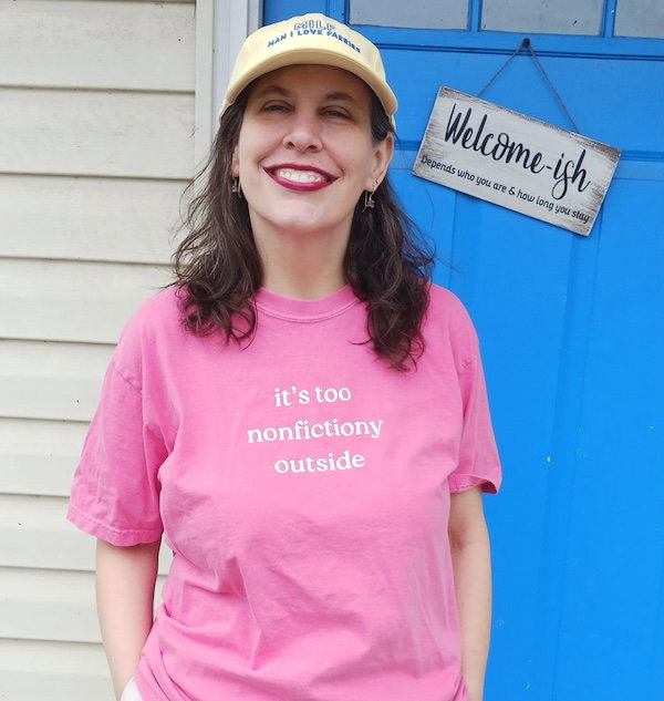 Crystal, a white woman with shoulder length brown hair, stands in a pink shirt that reads It's too nonfictiony outside, wearing a yellow ballcap that says MILF Man I Love Fairies. She is standing in front of a bright blue door with a sign that reads WELCOME-ISH Depends on who you are and how long you stay 